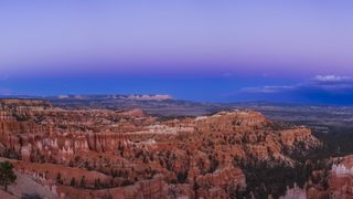 A stunning panoramic view of Bryce Canyon at twilight, showcasing vibrant orange and red rock formations known as hoodoos against a deepening purple and blue sky.