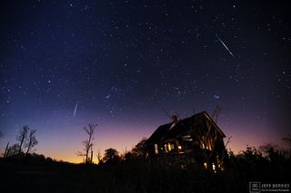 meteors streak through a star-studded sky with a house in the foreground.