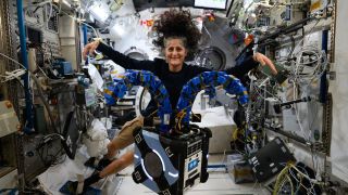 an astronaut in a black long sleeve shirt floats in a cluttered space station behind a cubical robot with two blue tentacles like bunny ears.