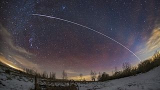 a bright white streak of light against a backdrop of stars. there is snow on the ground and a faint pink airglow in the air.